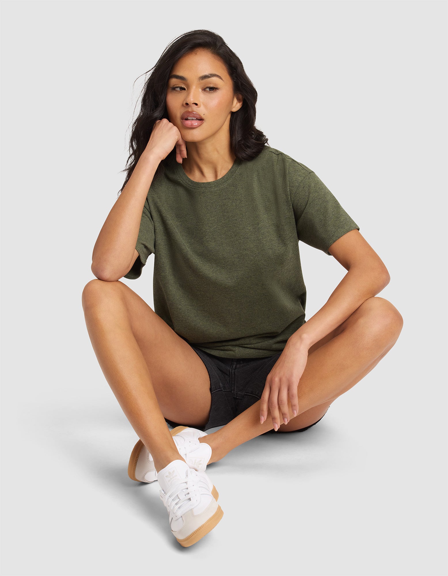 A woman with long dark hair, wearing Cozy Earth's Women's All Day Tee in olive green, black shorts, and white sneakers, sits cross-legged on a light gray floor, resting her chin on one hand and gazing slightly to the side. 