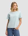 A woman with wavy blonde hair models the Cozy Earth Women's All Day Tee in light blue, paired with blue jeans. She stands against a plain light grey background, gazing slightly left with a relaxed expression.|Color:Tide