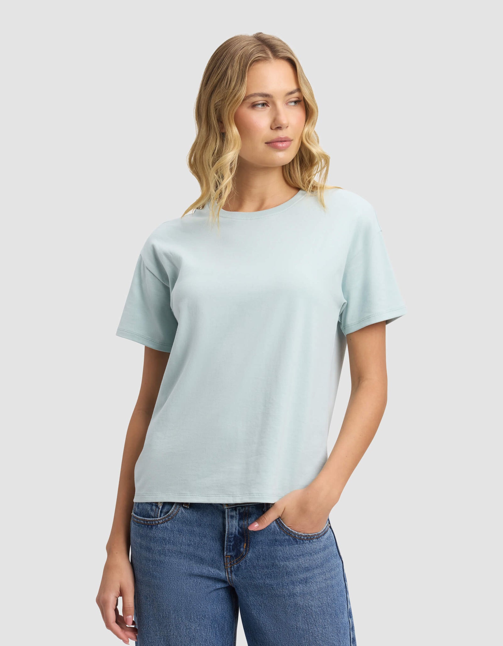 A woman with wavy blonde hair models the Cozy Earth Women's All Day Tee in light blue, paired with blue jeans. She stands against a plain light grey background, gazing slightly left with a relaxed expression.