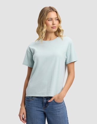 A woman with wavy blonde hair models the Cozy Earth Women's All Day Tee in light blue, paired with blue jeans. She stands against a plain light grey background, gazing slightly left with a relaxed expression.|Color:Tide