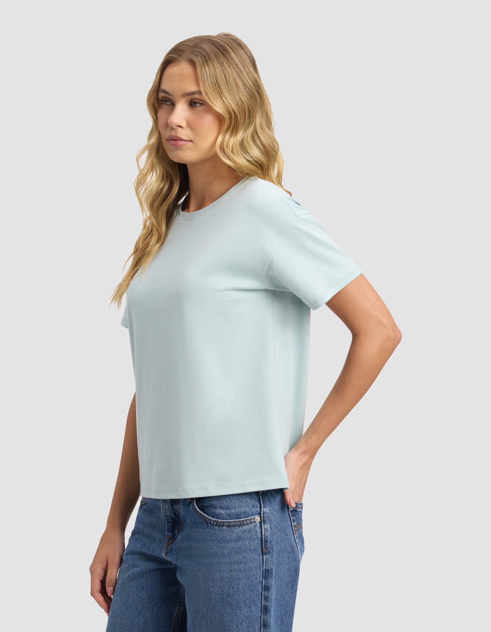 A woman with long blonde hair models the Cozy Earth Women's All Day Tee in light blue, paired with blue jeans, standing sideways with her left hand on her hip against a plain light gray background.