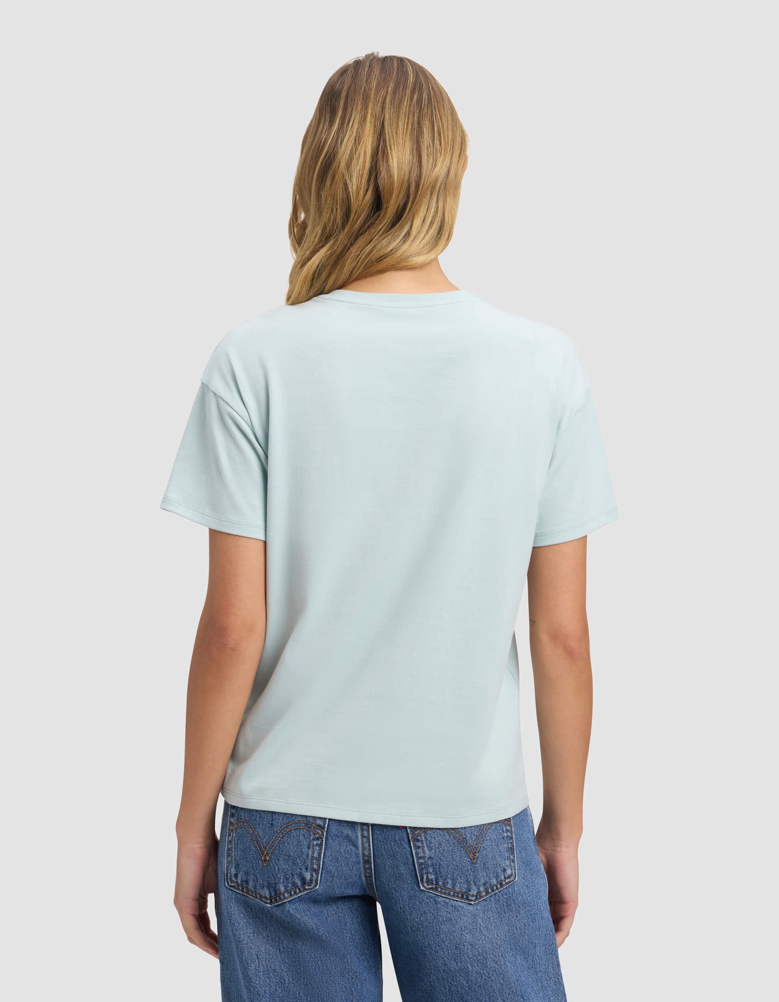 A woman with long blonde hair, dressed in Cozy Earth's Women's All Day Tee in light blue and blue jeans, faces away from the camera against a plain light gray background.