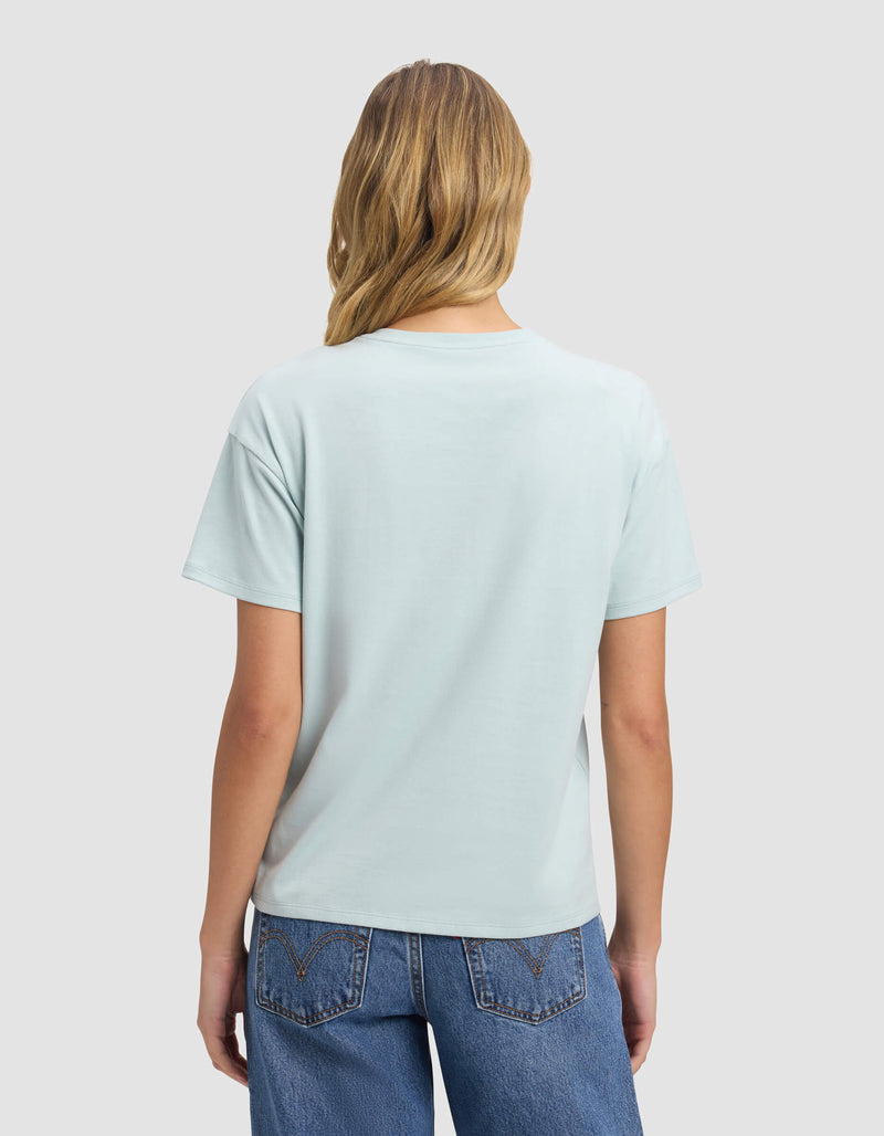 A woman with long blonde hair, dressed in Cozy Earth's Women's All Day Tee in light blue and blue jeans, faces away from the camera against a plain light gray background.