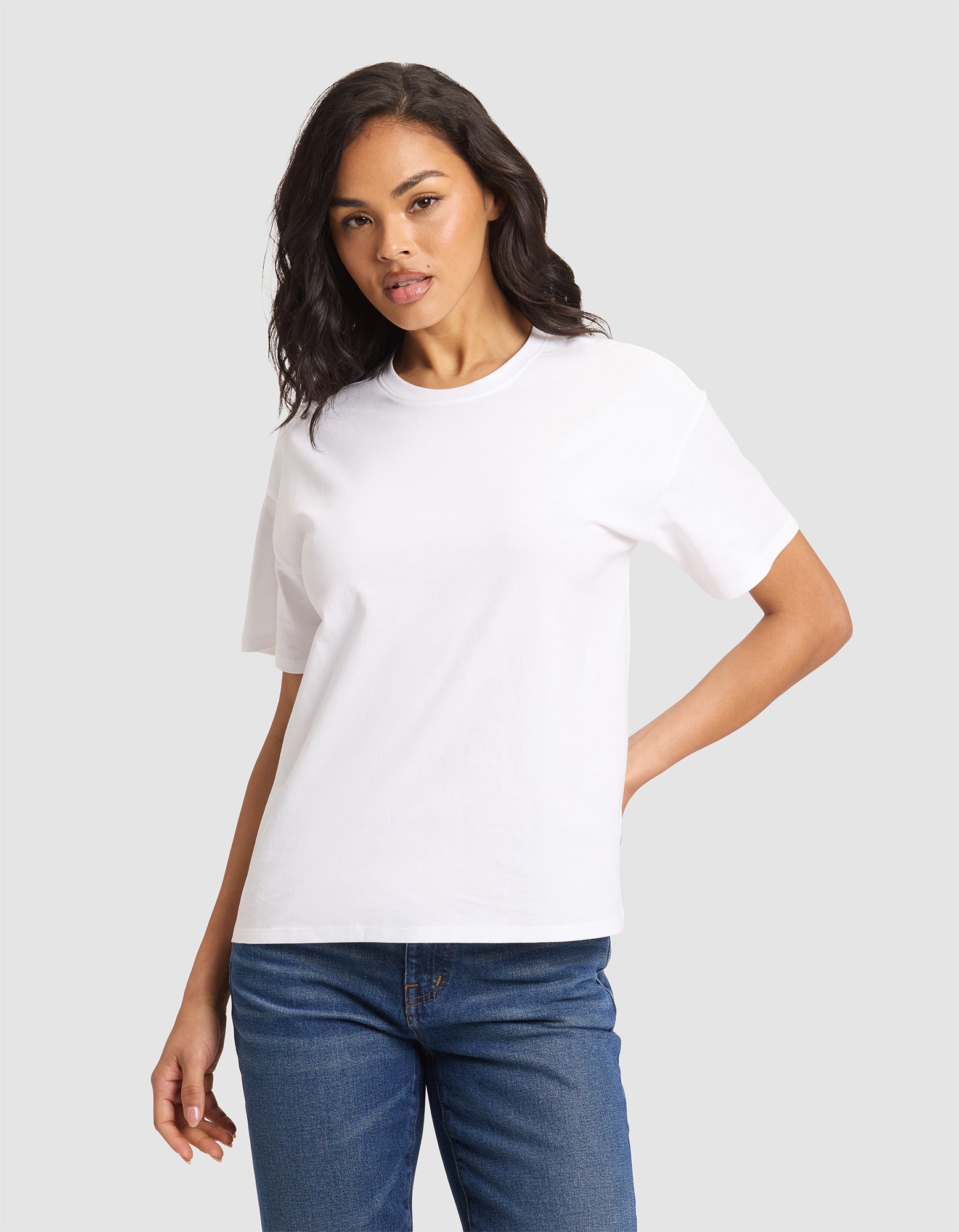 A woman models the Cozy Earth Women's All Day Tee in plain white, paired with blue jeans, standing against a light gray background with one hand on her hip and a neutral expression.  |Color:White