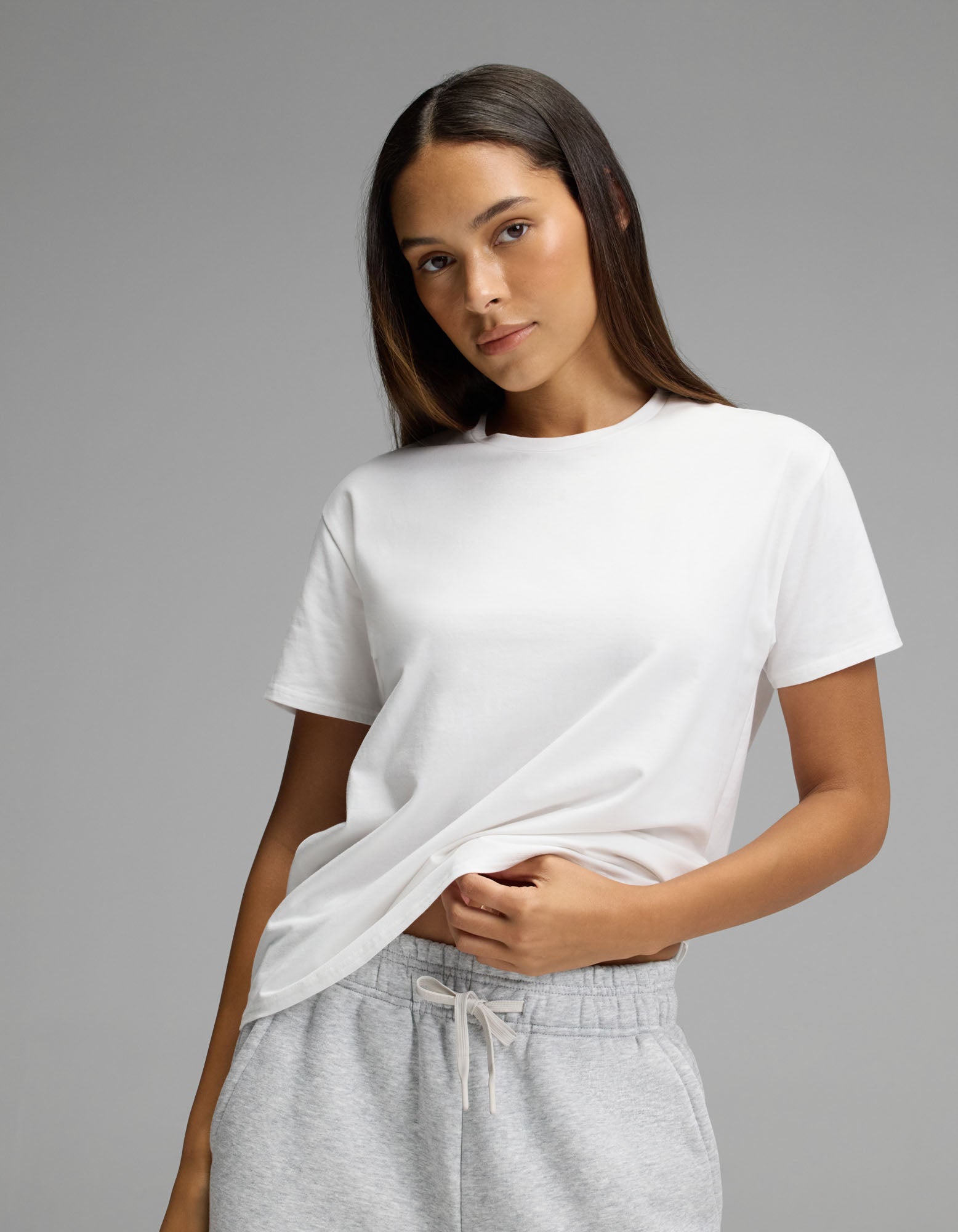 A woman wears the Cozy Earth Women’s All Day Tee in plain white and light gray sweatpants, standing against a neutral background. She looks at the camera with a slight smile while holding the hem of her shirt.|Color:White