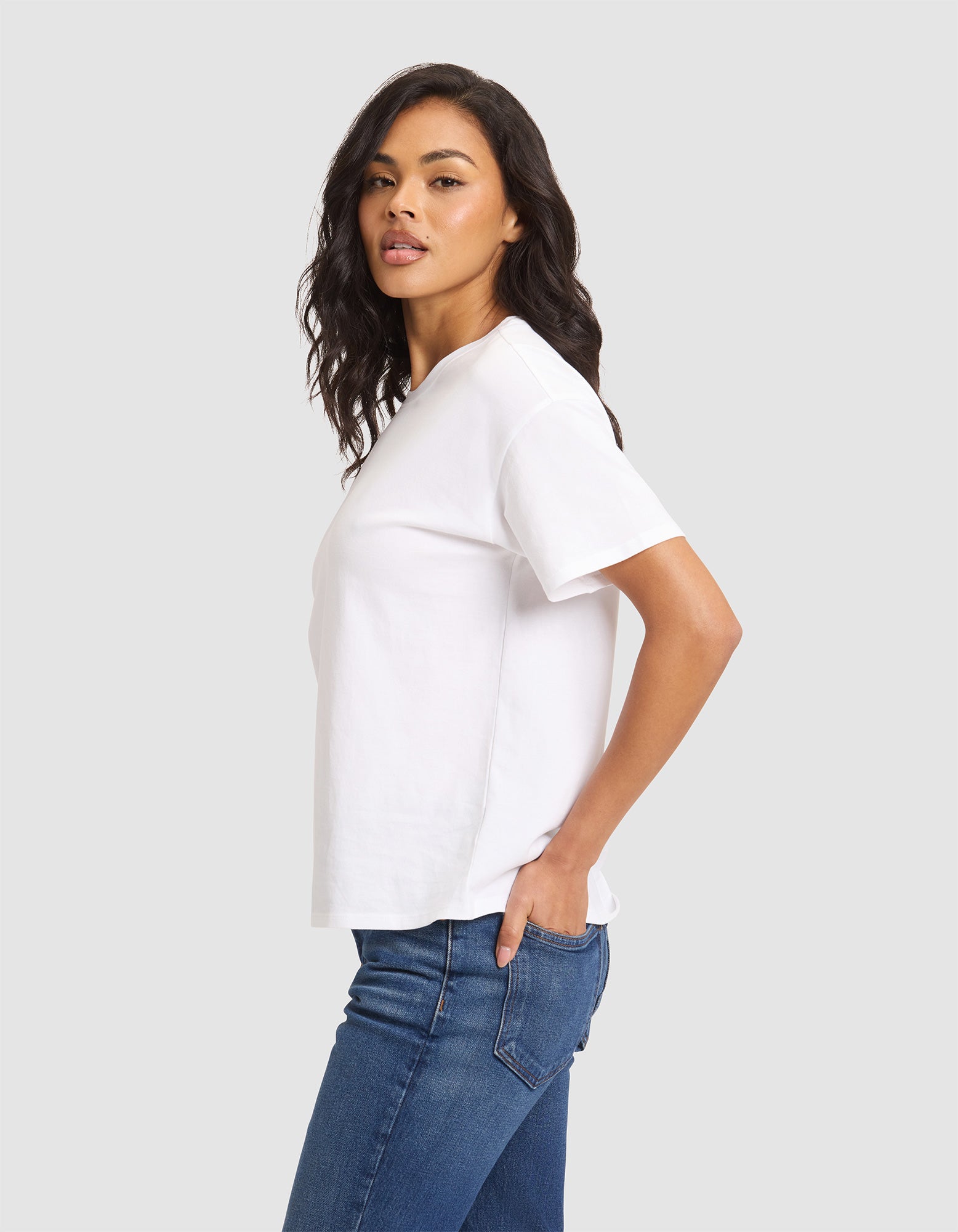 A woman with long, wavy dark hair stands sideways against a light gray background, wearing the Cozy Earth Women's All Day Tee in white and blue jeans, with her hand in her pocket and looking slightly toward the camera.