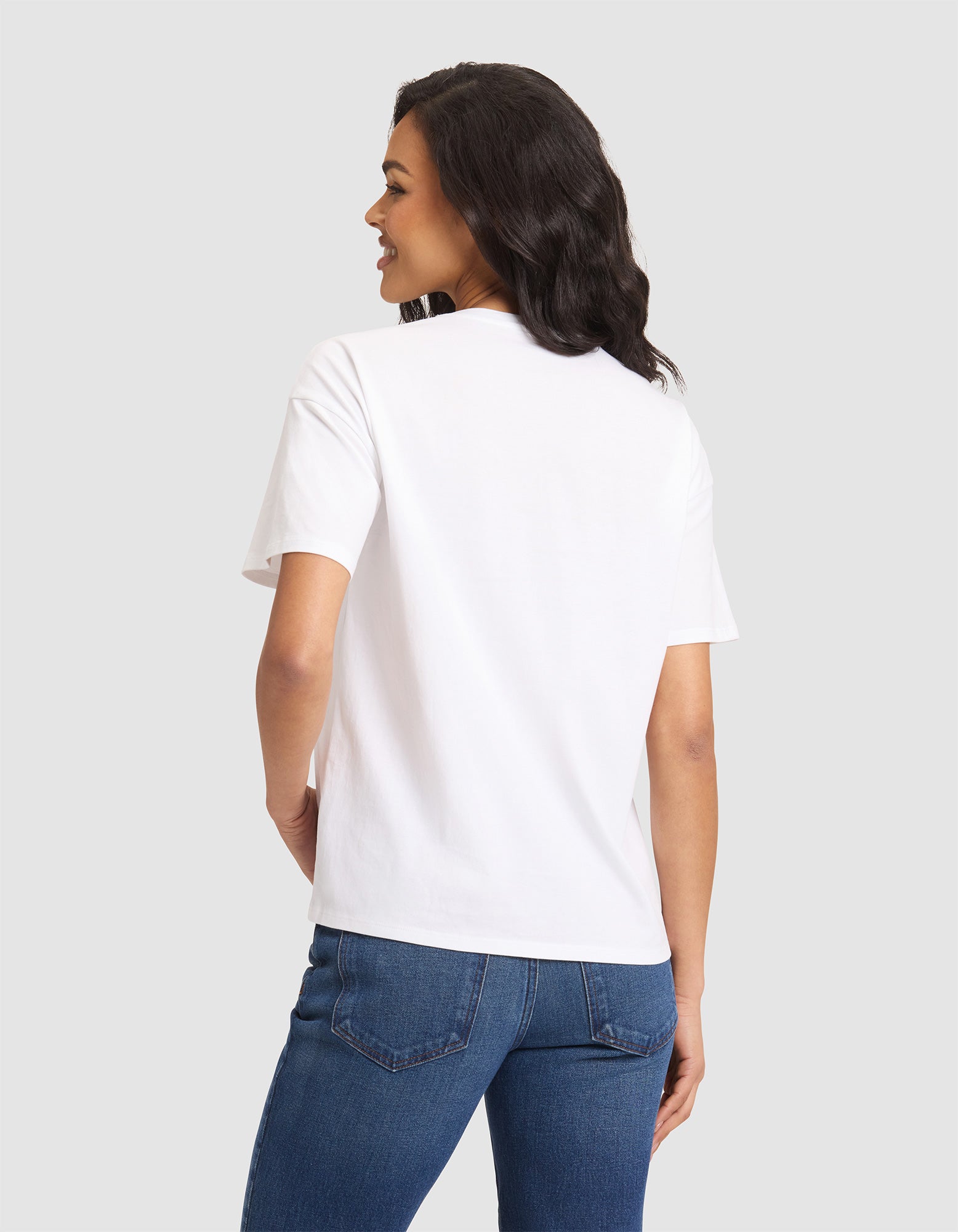A woman with shoulder-length dark hair models the Cozy Earth Women's All Day Tee, a plain white short-sleeve T-shirt, paired with blue jeans. She stands facing away on a light gray background, smiling with her left hand on her hip. 