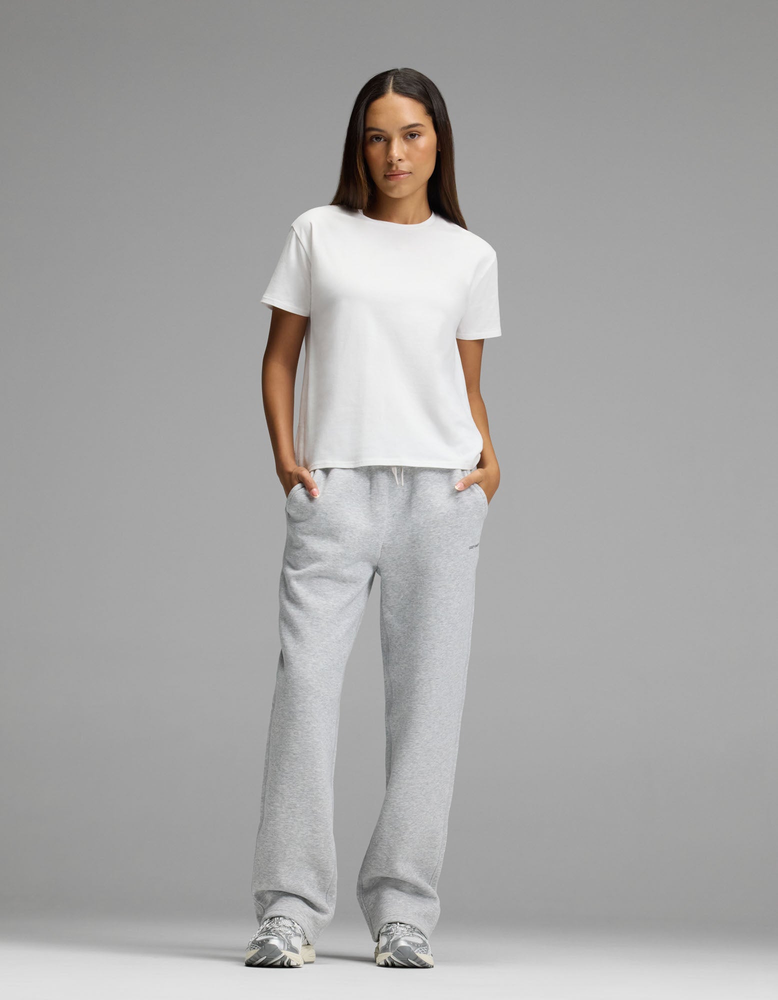 A woman stands facing forward against a plain gray background, wearing the Cozy Earth Women’s All Day Tee in white, light gray sweatpants, and sneakers, with her hands in her pockets.