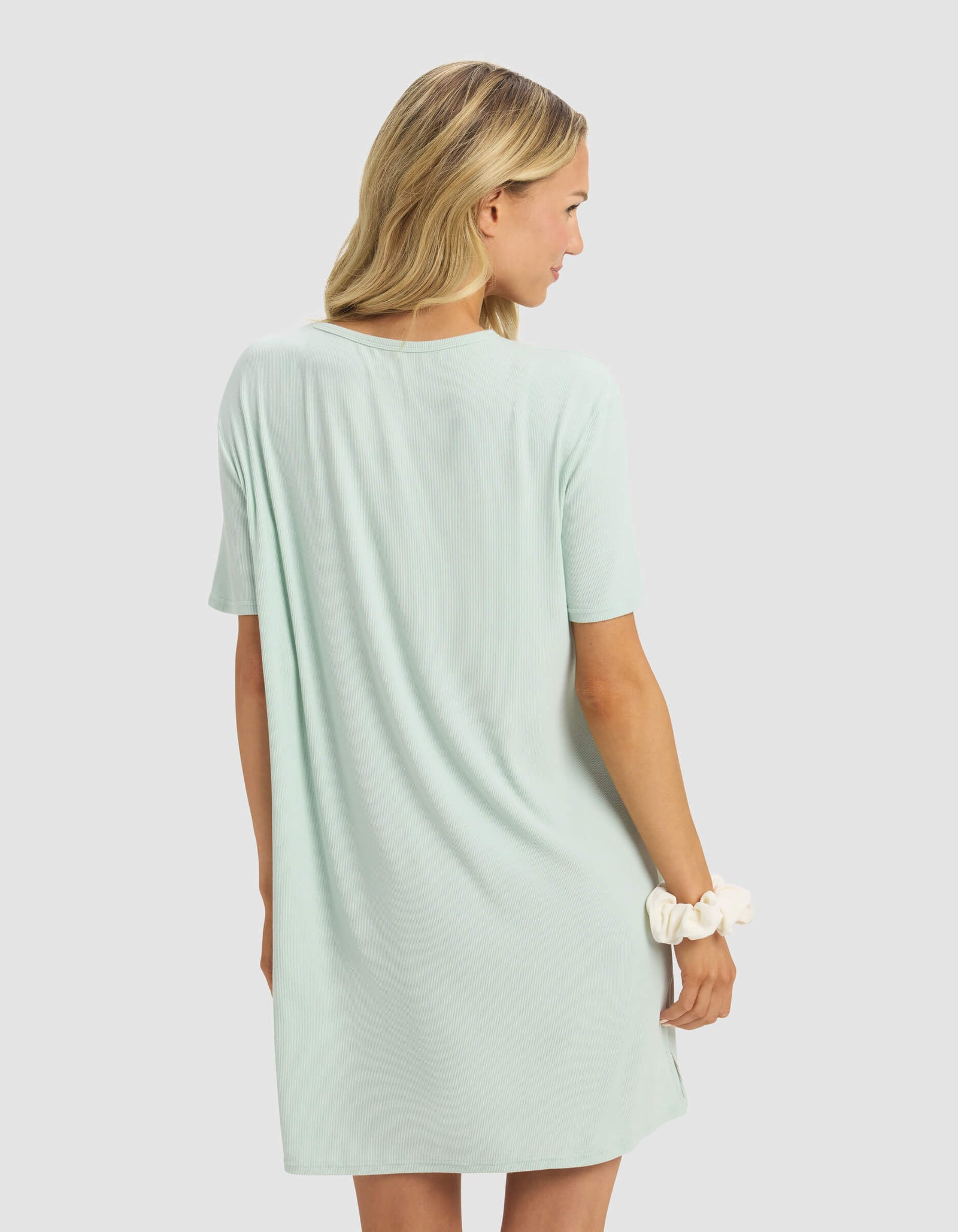 A blonde woman models the Cozy Earth Women's Bamboo Rib-Knit Boyfriend Dress in light mint green, short sleeves. She stands against a plain light gray background, facing away and holding a white scrunchie in her right hand.