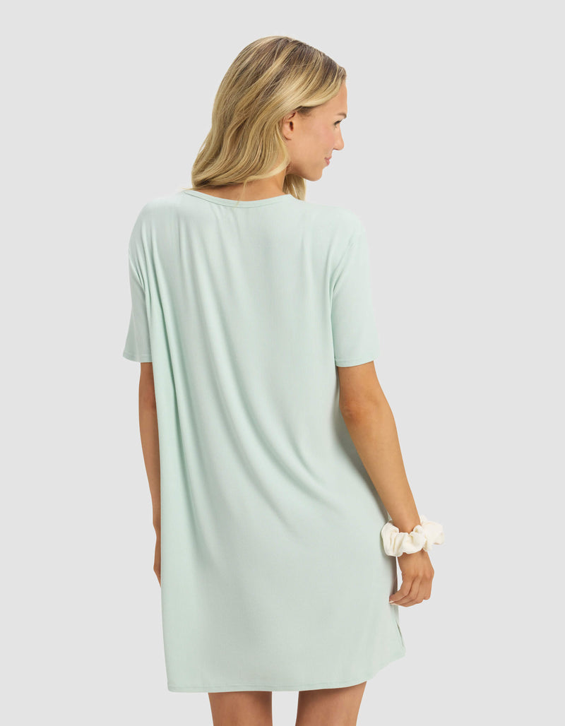 A blonde woman models the Cozy Earth Women's Bamboo Rib-Knit Boyfriend Dress in light mint green, short sleeves. She stands against a plain light gray background, facing away and holding a white scrunchie in her right hand.