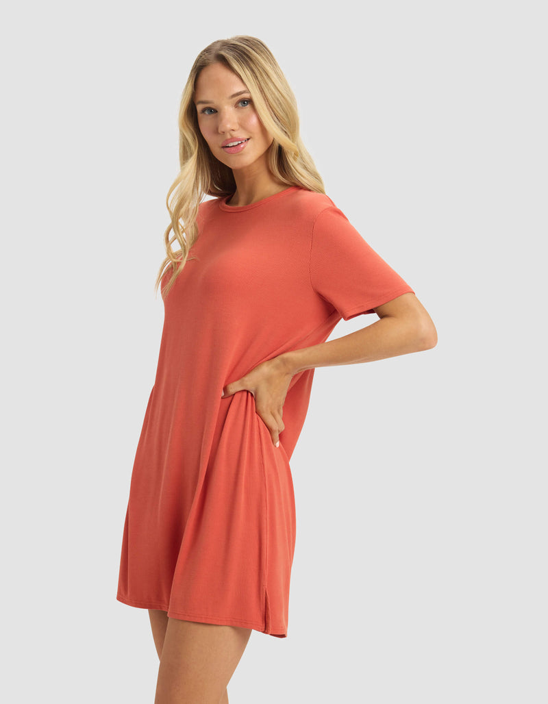 A woman with long blonde hair, wearing the Cozy Earth Women's Bamboo Rib-Knit Boyfriend Dress in coral, poses with one hand on her hip and a slight smile against a plain light gray background.