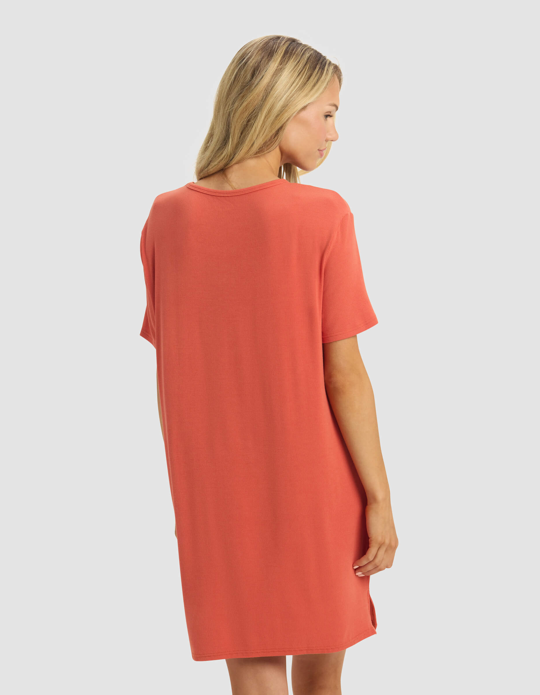 A woman with blonde hair, facing away, wears the Cozy Earth Women's Bamboo Rib-Knit Boyfriend Dress in coral—a loose-fitting, short-sleeve style falling just above the knee. The background is plain light gray.