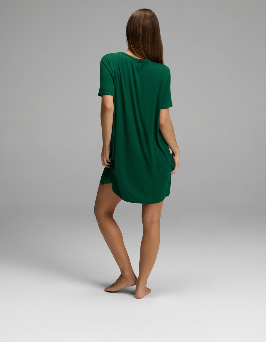 A person with long brown hair stands barefoot, facing away, wearing the Cozy Earth Women’s Bamboo Rib-Knit Boyfriend Sleep Dress in green. The background is plain and light gray.