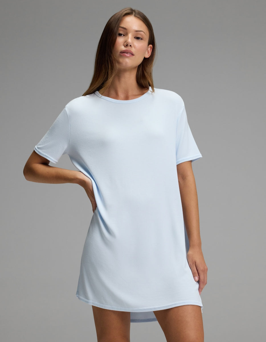 A woman models the Cozy Earth Women’s Bamboo Rib-Knit Boyfriend Sleep Dress in light blue, standing against a plain gray background with one hand on her hip and the other relaxed by her side.