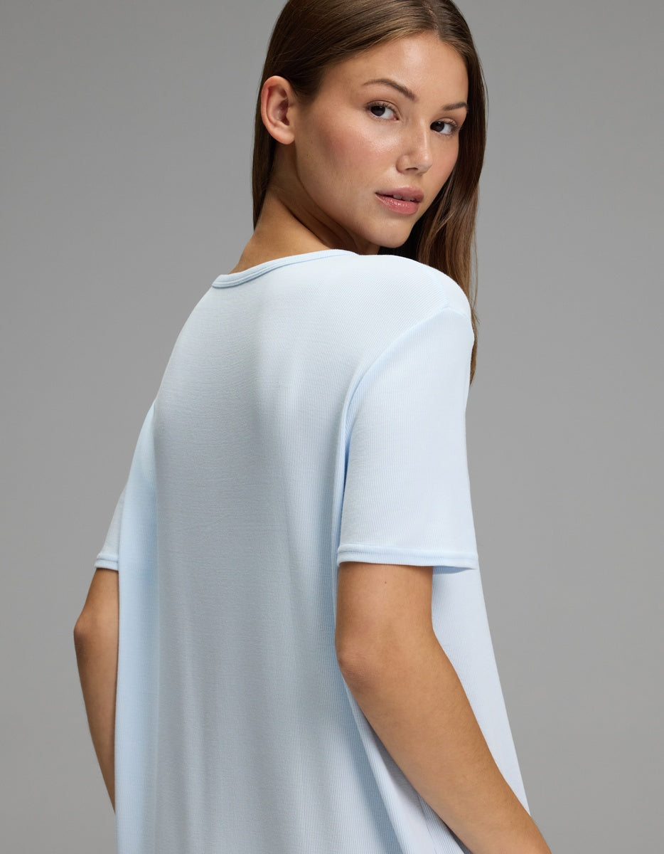 A young woman with light brown hair, wearing the Cozy Earth Women’s Bamboo Rib-Knit Boyfriend Sleep Dress in light blue, looks over her shoulder against a plain gray background.