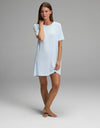 A woman stands barefoot on a plain background, wearing the Cozy Earth Women’s Bamboo Rib-Knit Boyfriend Sleep Dress in light blue. One hand touches her face while the other rests by her side.|Color:Frost