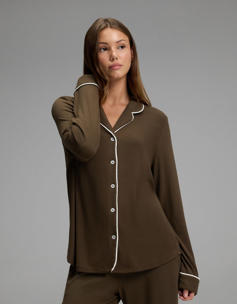 A woman wearing the Cozy Earth Women’s Bamboo Rib-Knit Classic Long Sleeve Pajama Top in brown with white piping stands against a plain gray background, looking to the side with one hand touching her neck.|Color:Espresso