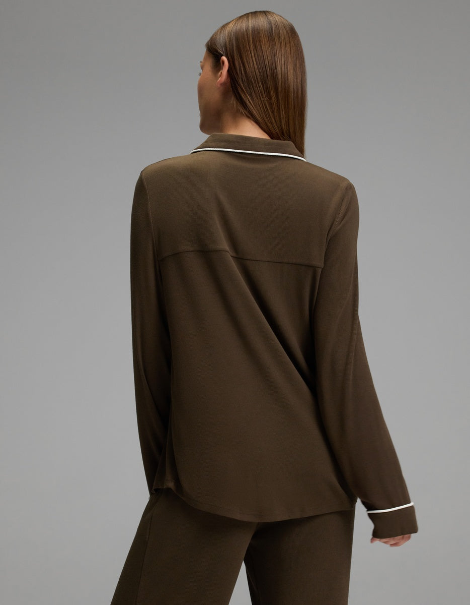 A person with straight brown hair is seen from behind wearing a Cozy Earth Women’s Bamboo Rib-Knit Classic Long Sleeve Pajama Top in brown, featuring white piping along the collar and cuffs, paired with matching pants. The background is plain and gray.