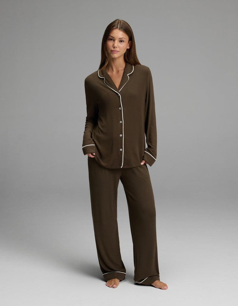 A woman stands barefoot on a gray background, looking at the camera with a neutral expression while wearing the Cozy Earth Women’s Bamboo Rib-Knit Classic Long Sleeve Pajama Top in dark brown with white trim.