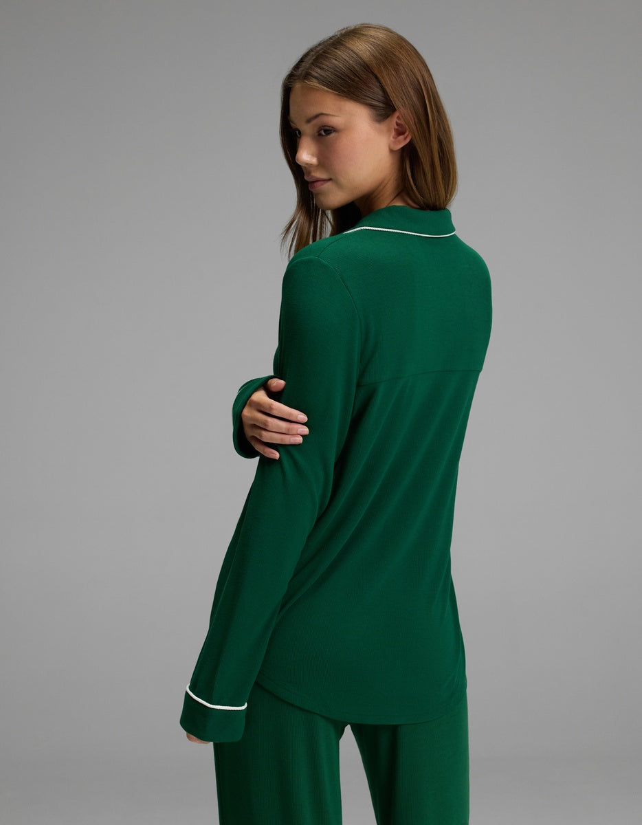 A woman with long brown hair stands and looks over her shoulder against a plain gray background, wearing the Cozy Earth Women’s Bamboo Rib-Knit Classic Long Sleeve Pajama Top in dark green with white trim.