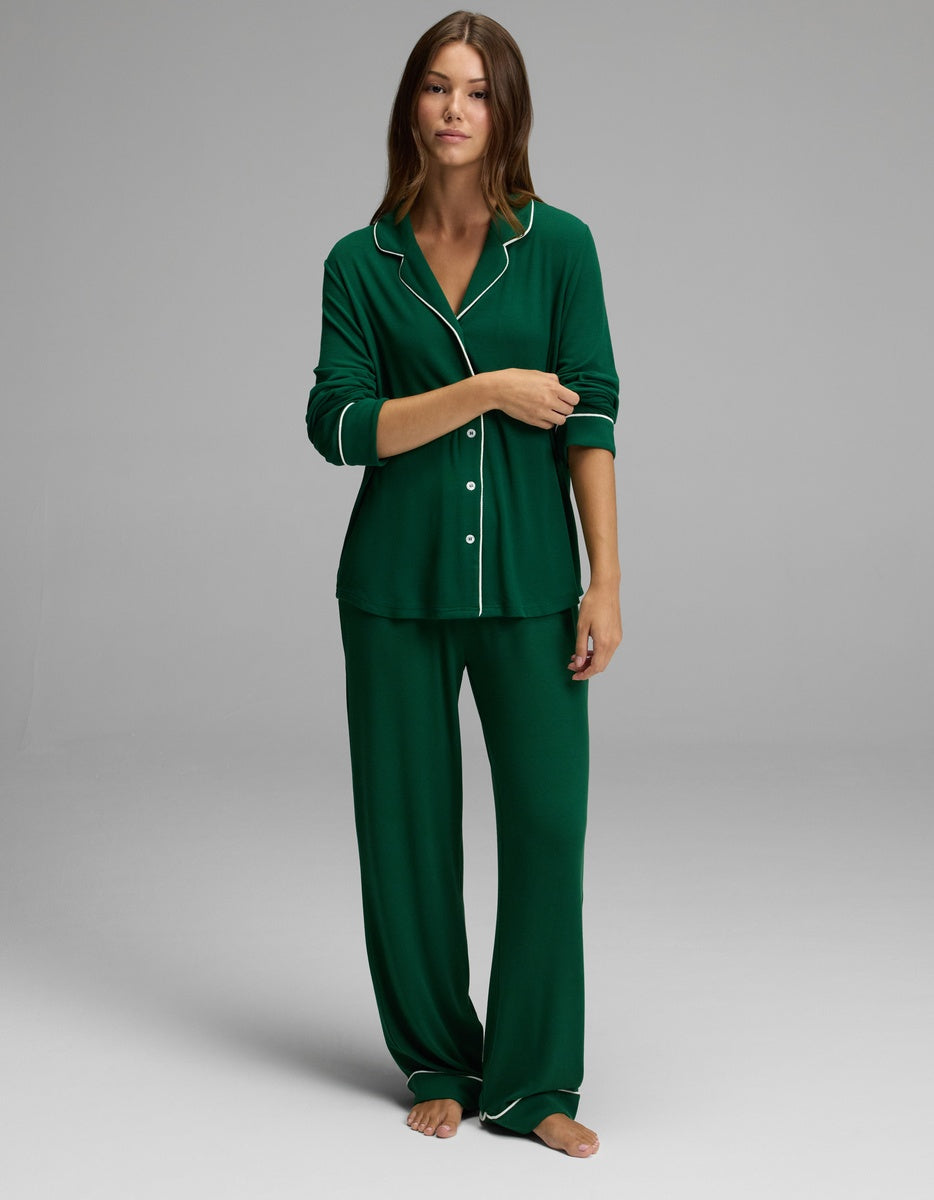 A woman stands barefoot against a plain light gray background, wearing the Cozy Earth Women’s Bamboo Rib-Knit Classic Long Sleeve Pajama Top in dark green with white piping, paired with matching loose pants.