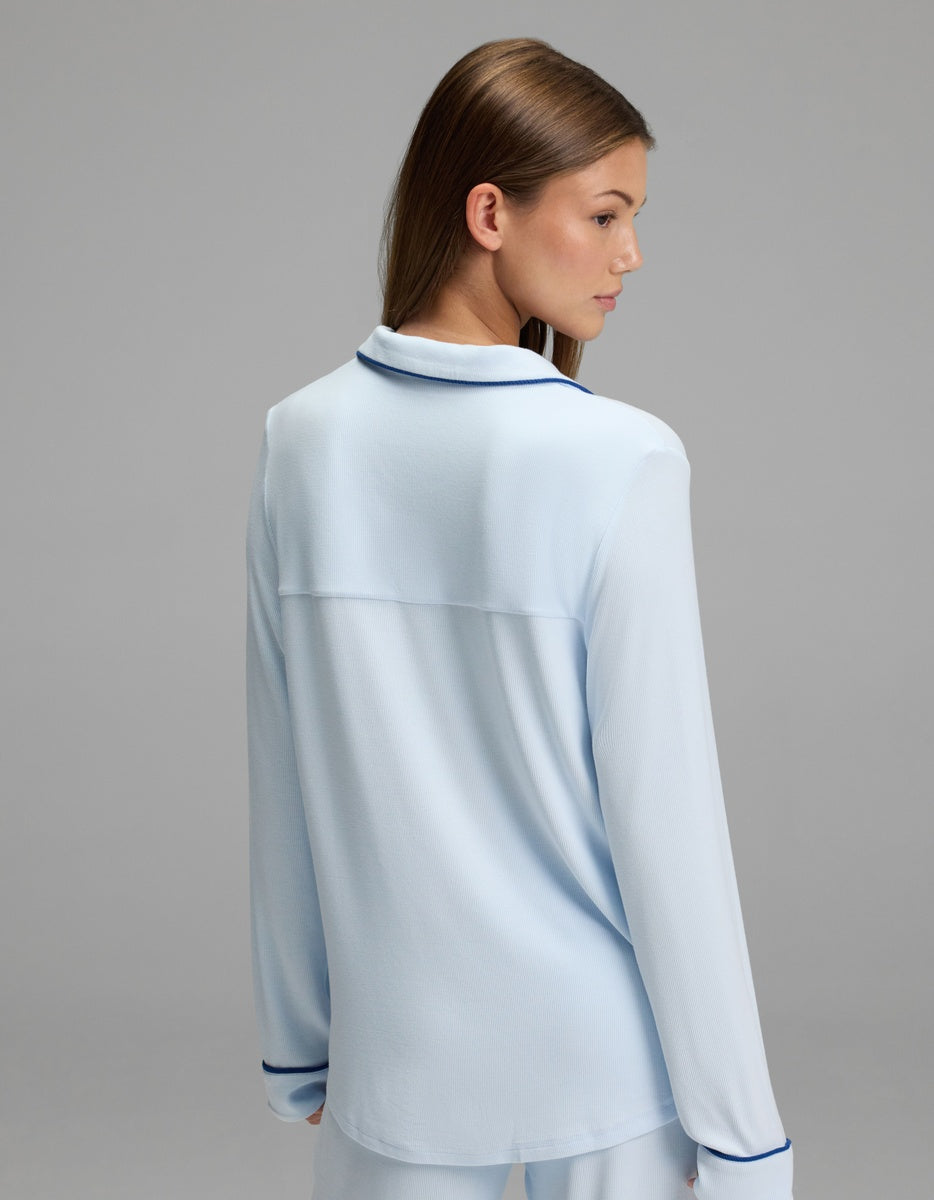 A woman with straight brown hair is standing and facing away, wearing the Cozy Earth Women’s Bamboo Rib-Knit Classic Long Sleeve Pajama Top in light blue with dark piping on the collar and cuffs, against a plain gray background.