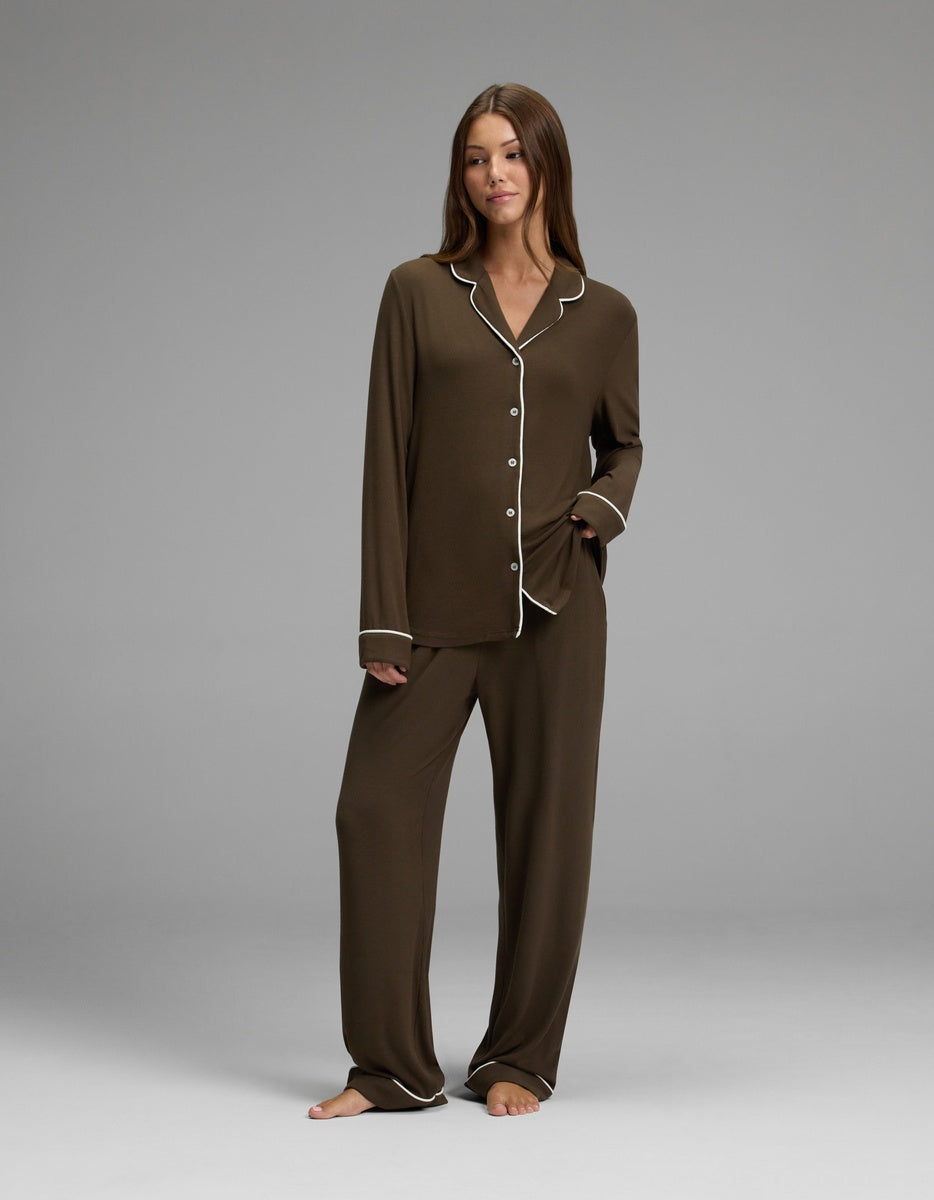 A woman stands barefoot, wearing the Cozy Earth Women’s Bamboo Rib-Knit Classic Long Sleeve Pajama Set—loose brown pajamas with white trim, featuring a button-up top and matching pants—as she poses against a plain gray background.|Color:Espresso
