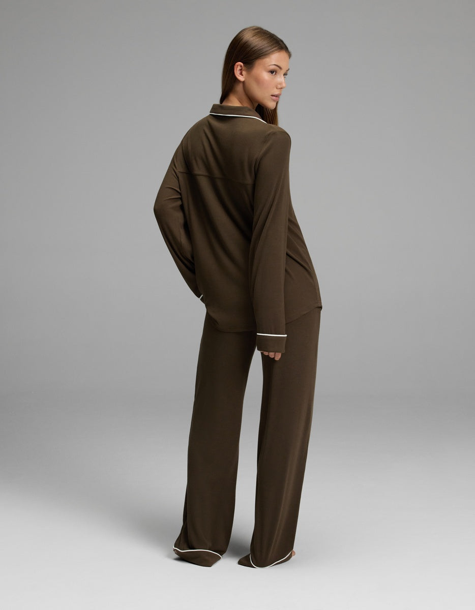 A woman stands in a studio, facing away but looking over her shoulder, wearing the Cozy Earth Women’s Bamboo Rib-Knit Classic Long Sleeve Pajama Set in a loose-fitting brown style with white trim. The background is plain and gray.