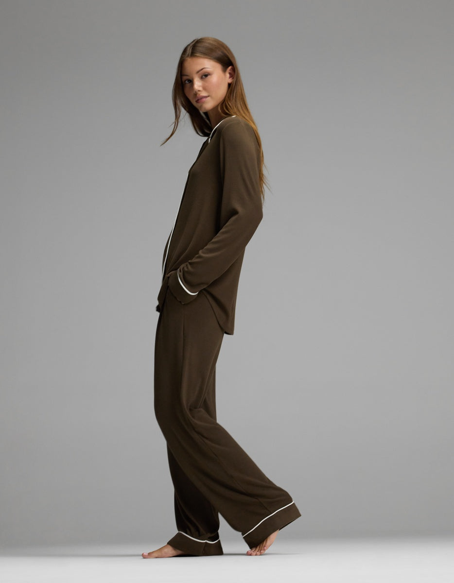 A woman stands barefoot in profile against a plain gray background, wearing the Cozy Earth Women’s Bamboo Rib-Knit Classic Long Sleeve Pajama Set, featuring a matching brown color with white piping on the collar, cuffs, and hems.|Color:Espresso