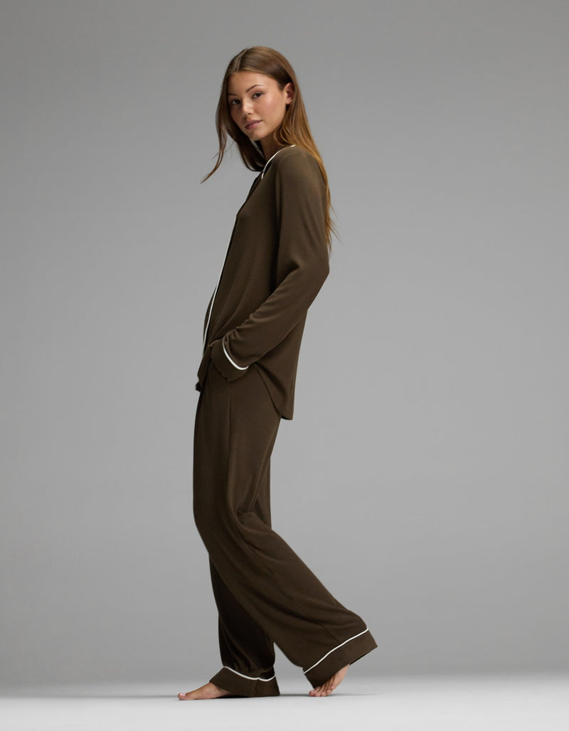 A woman stands barefoot in profile against a plain gray background, wearing the Cozy Earth Women’s Bamboo Rib-Knit Classic Long Sleeve Pajama Set, featuring a matching brown color with white piping on the collar, cuffs, and hems.
