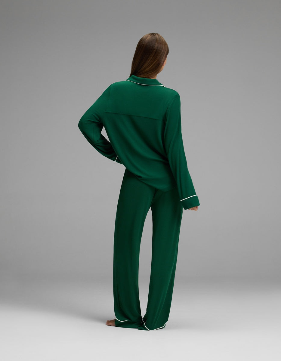 A person stands barefoot on a gray background, facing away. They are wearing Cozy Earth’s Women’s Bamboo Rib-Knit Classic Long Sleeve Pajama Set in dark green with white trim on the cuffs and pants. Their right hand rests on their hip.