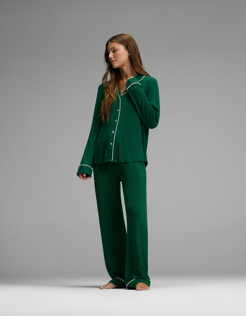 A woman stands barefoot, wearing Cozy Earth’s Women’s Bamboo Rib-Knit Classic Long Sleeve Pajama Set in dark green with white trim. The set features a long-sleeve button-up top and wide-leg pants. She gazes to her left against a plain gray background.