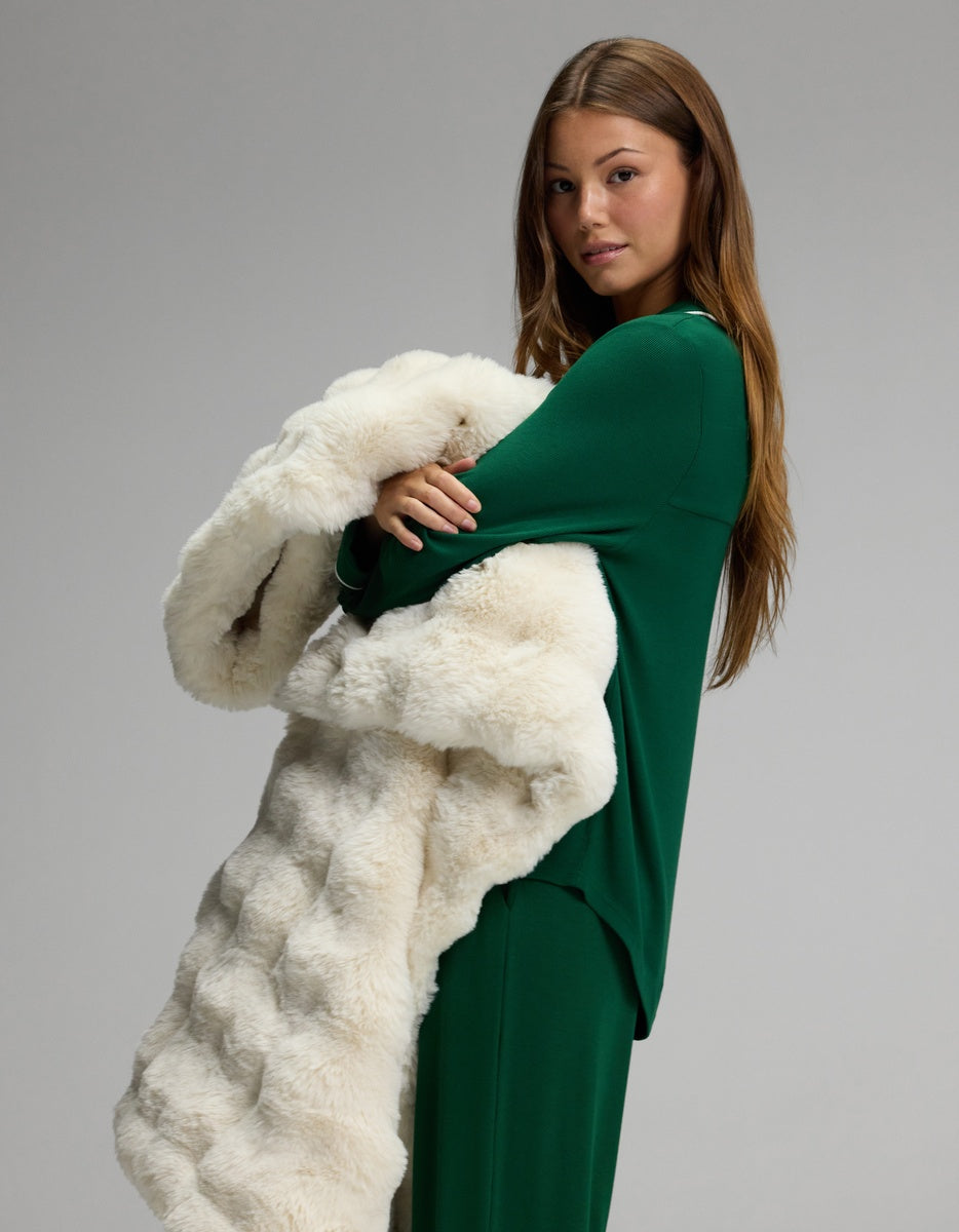 A woman wearing the Cozy Earth Women’s Bamboo Rib-Knit Classic Long Sleeve Pajama Set in green stands against a plain gray background, holding a large, fluffy white fur coat draped over her arms.