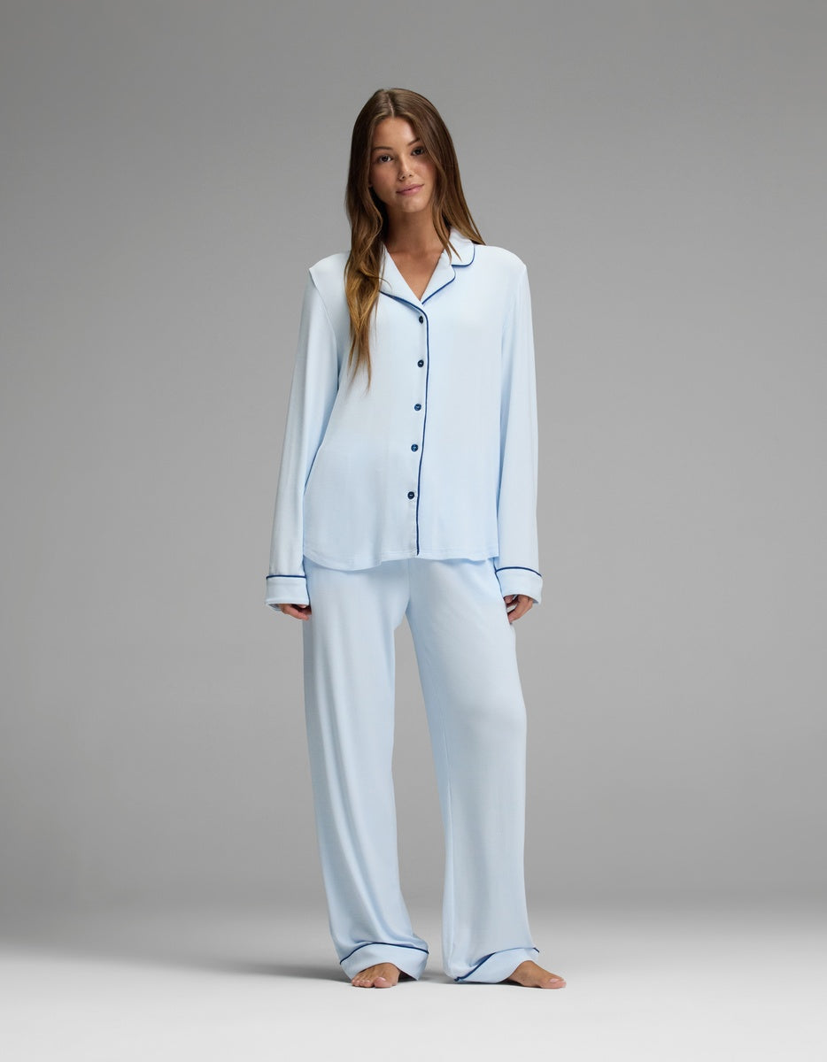A woman stands barefoot against a plain gray background wearing the Cozy Earth Women’s Bamboo Rib-Knit Classic Long Sleeve Pajama Set, featuring loose, light blue pants and a matching button-up top with dark trims.
