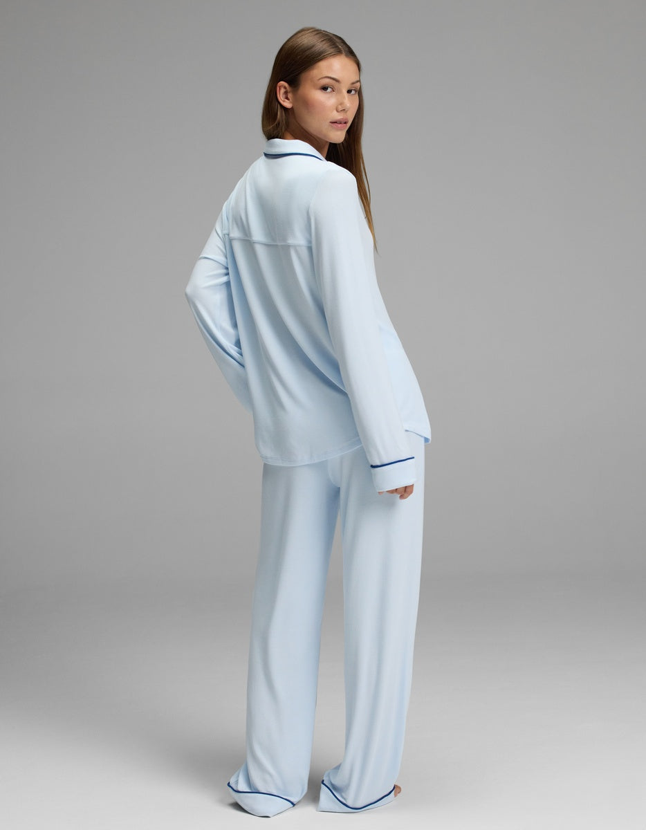 A woman models the Cozy Earth Women’s Bamboo Rib-Knit Classic Long Sleeve Pajama Set in light blue with dark trim, standing against a plain gray background and glancing over her shoulder.