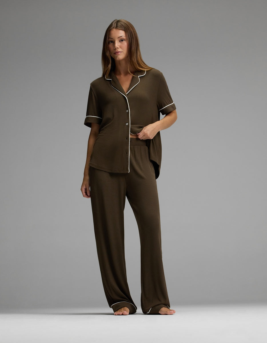 A woman stands barefoot against a plain gray background, wearing the Cozy Earth Women’s Bamboo Rib-Knit Classic Pajama Pant in brown with white trim, paired with a matching short-sleeve button-up top and loose pants.