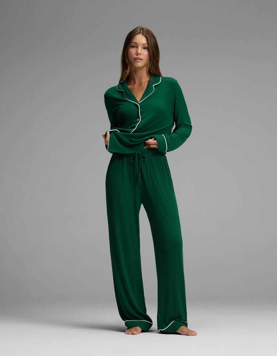 A woman stands barefoot against a plain background, wearing Cozy Earth’s Women’s Bamboo Rib-Knit Classic Pajama Pant in dark green with white piping and long sleeves. She looks at the camera with a relaxed expression, her hands tucked into the pockets of her pajama pants.