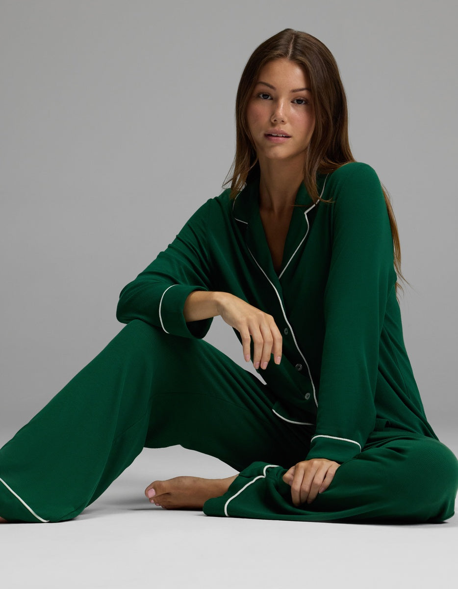 A woman with long brown hair sits barefoot on the floor, wearing Cozy Earth’s Women’s Bamboo Rib-Knit Classic Pajama Pant in dark green with white piping, against a plain gray background.