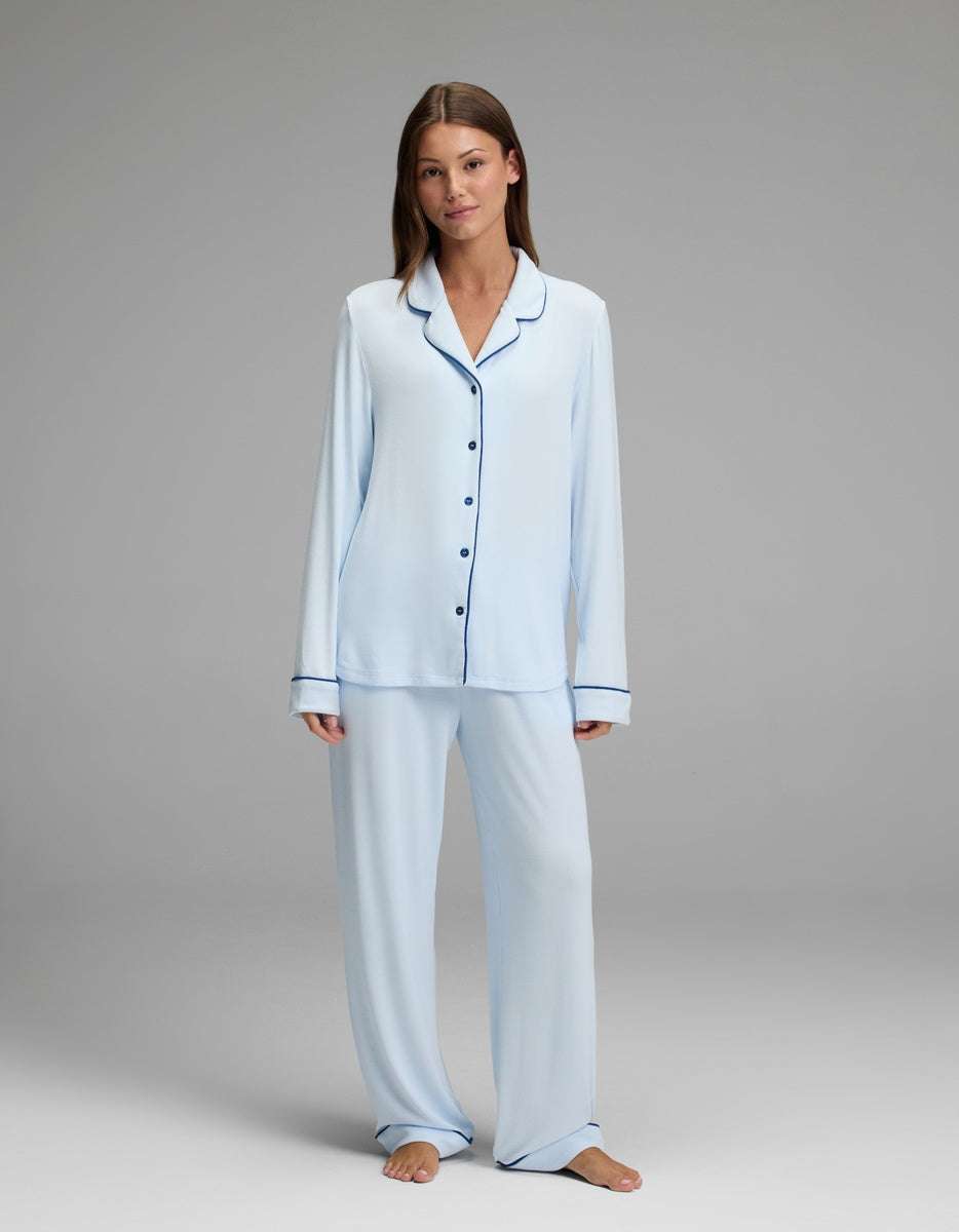 A woman stands barefoot against a plain gray background, wearing the Cozy Earth Women’s Bamboo Rib-Knit Classic Pajama Pant in light blue and a matching long-sleeve button-up pajama top with dark trim.