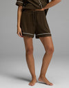 A person is wearing the Cozy Earth Women’s Bamboo Rib-Knit Classic Pajama Short in dark brown with white piping, paired with a matching pajama top. They are barefoot and standing on a plain light gray background, with only their torso and legs visible.|Color:Espresso