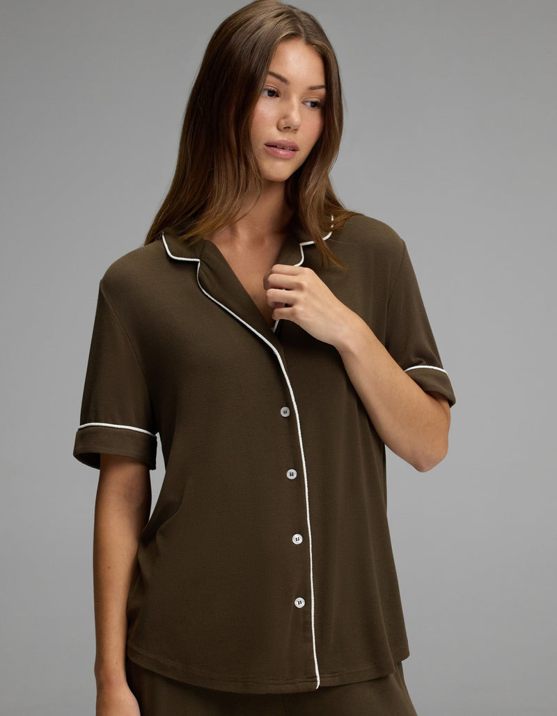 A woman with long brown hair wears the Cozy Earth Women’s Bamboo Rib-Knit Classic Short Sleeve Pajama Top, featuring dark brown fabric with white piping and buttons, while standing against a plain gray background.