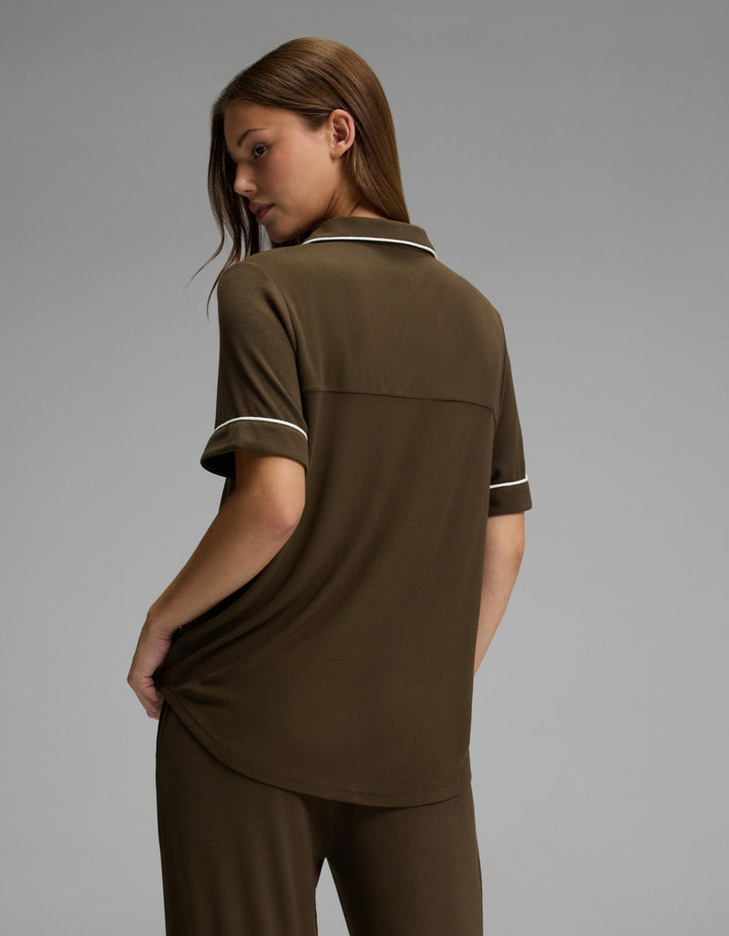 A woman stands facing away, wearing the Cozy Earth Women’s Bamboo Rib-Knit Classic Short Sleeve Pajama Top in brown with matching pants featuring white trim, set against a plain gray background.