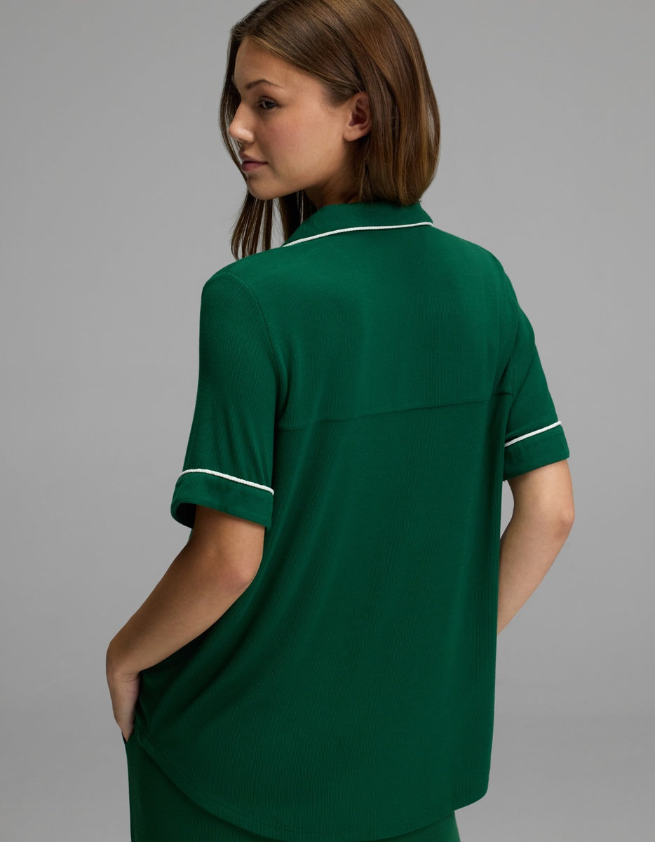 A woman with brown hair wears the Cozy Earth Women’s Bamboo Rib-Knit Classic Short Sleeve Pajama Top, a green collared shirt with white trim, as she stands against a plain gray background with her back mostly to the camera, head turned slightly to the side and hands in her pockets.