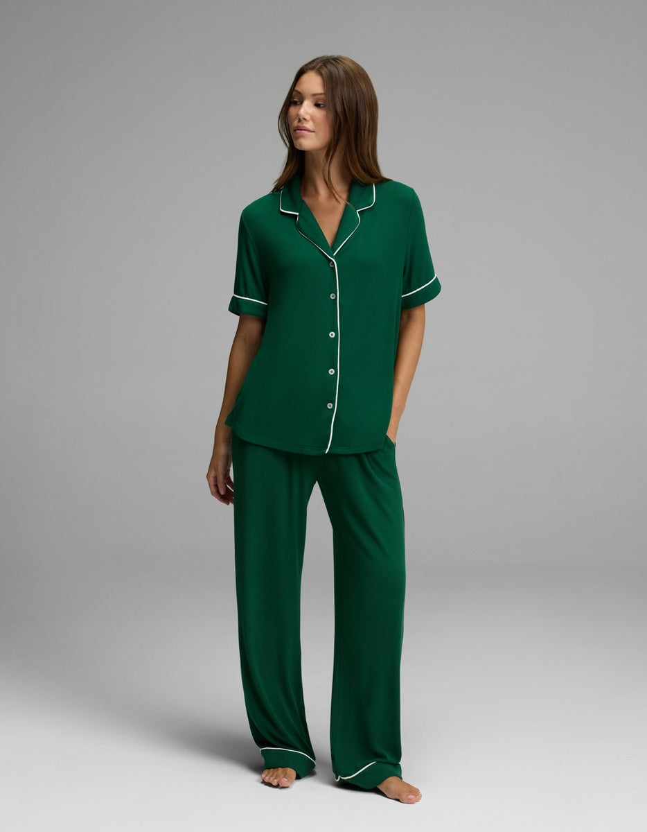 A woman stands barefoot wearing the Cozy Earth Women’s Bamboo Rib-Knit Classic Short Sleeve Pajama Top in green with white trim. She has straight brown hair and is looking slightly to the side, set against a plain gray background.