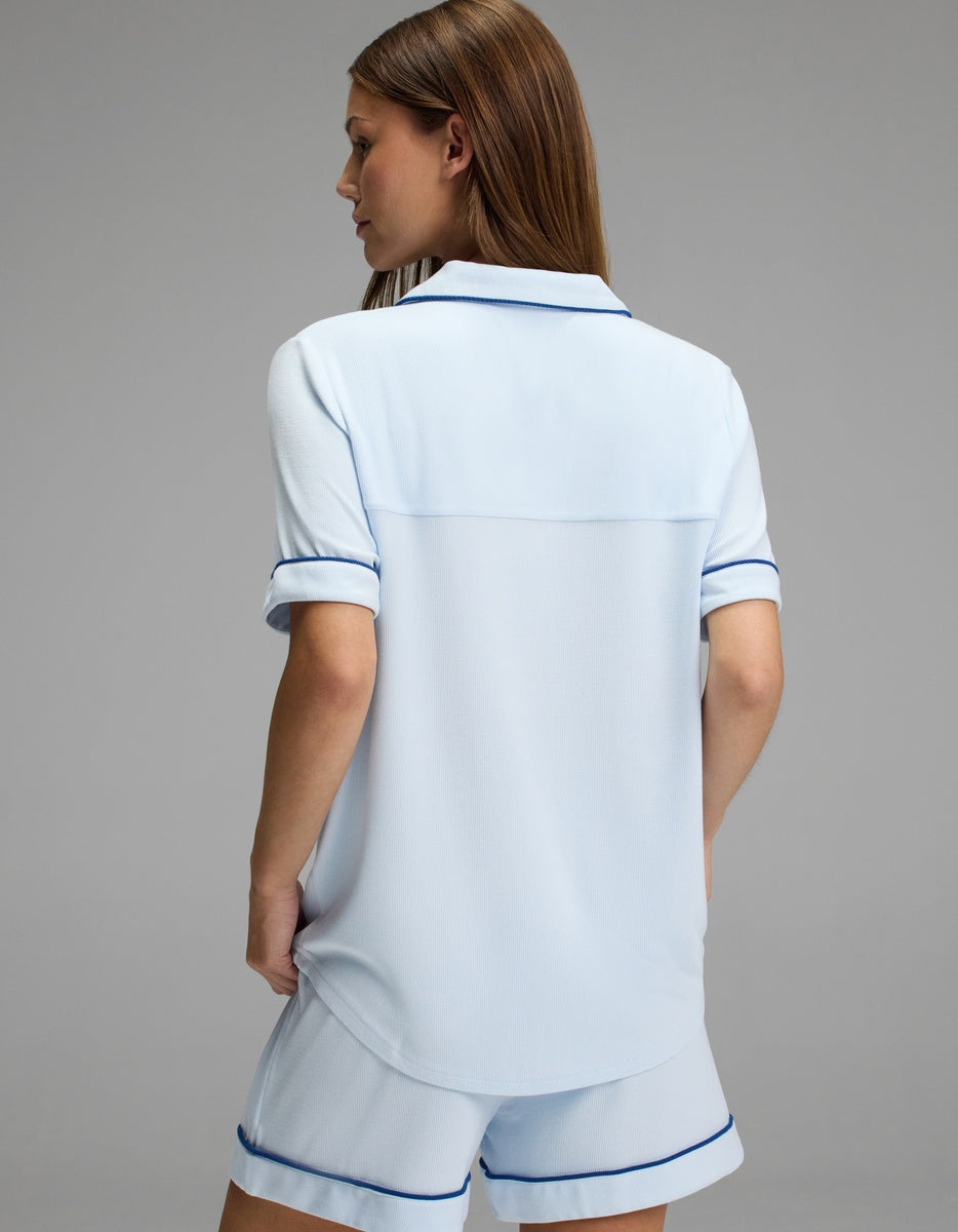 A person with long brown hair is shown from the back, wearing the Cozy Earth Women’s Bamboo Rib-Knit Classic Short Sleeve Pajama Top in light blue with dark blue trim, standing against a plain gray background.
