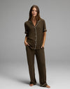 A woman stands barefoot, wearing the Cozy Earth Women’s Bamboo Rib-Knit Classic Short Sleeve & Pant Pajama Set in a loose-fitting, dark brown style with white piping. The set features a short-sleeve, button-up top and long pants. She is posed against a plain gray background.|Color:Espresso