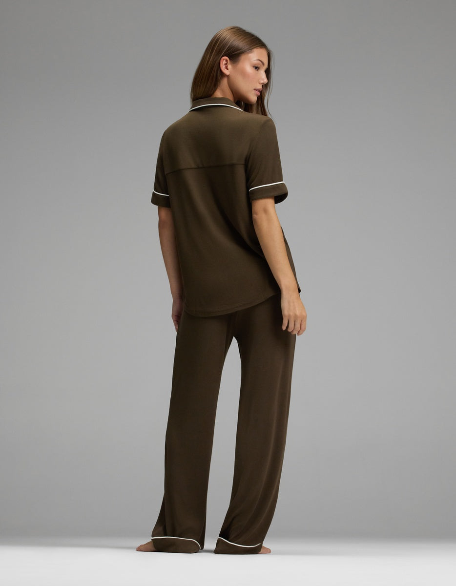 A woman stands barefoot, facing away, modeling the Cozy Earth Women’s Bamboo Rib-Knit Classic Short Sleeve & Pant Pajama Set in dark brown with white trim on the sleeves, collar, and pant cuffs, against a plain gray background.|Color:Espresso