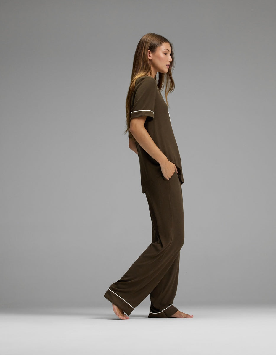 A woman with long hair stands barefoot in profile, wearing the Cozy Earth Women’s Bamboo Rib-Knit Classic Short Sleeve & Pant Pajama Set—a loose-fitting brown ensemble with white trim—against a plain gray background.