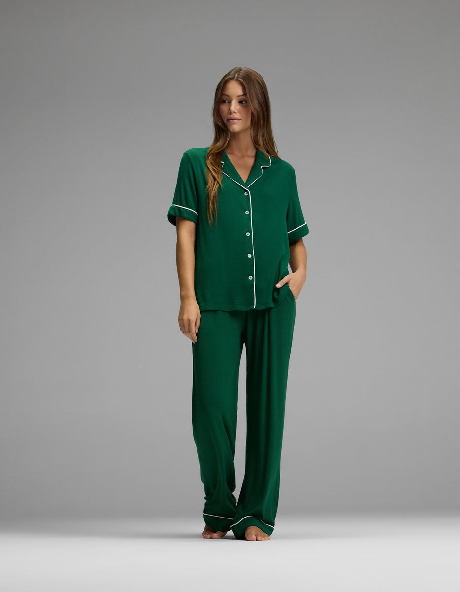 A woman stands barefoot against a plain gray background, wearing the Cozy Earth Women’s Bamboo Rib-Knit Classic Short Sleeve & Pant Pajama Set in green with white piping. The pajama set features a short-sleeve button-up top and loose-fitting pants.