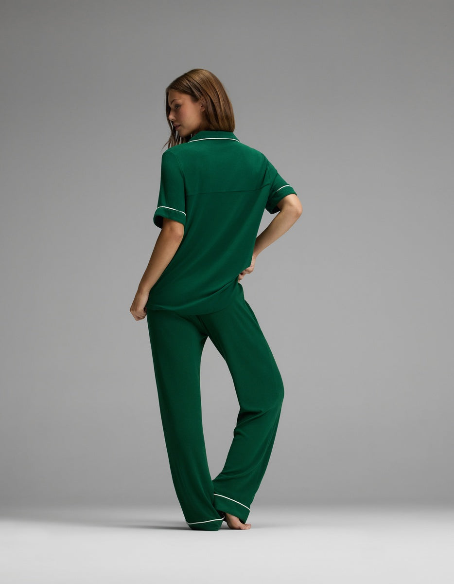 A woman stands with her back to the camera, wearing the Cozy Earth Women’s Bamboo Rib-Knit Classic Short Sleeve & Pant Pajama Set in green with white trim, set against a plain studio backdrop.